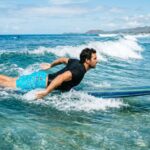 A male surfer wearing bright blue board shorts paddling on a surfboard in clear ocean waves